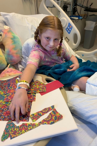 child smiling and posing with their artwork, which features bright patterned paper 